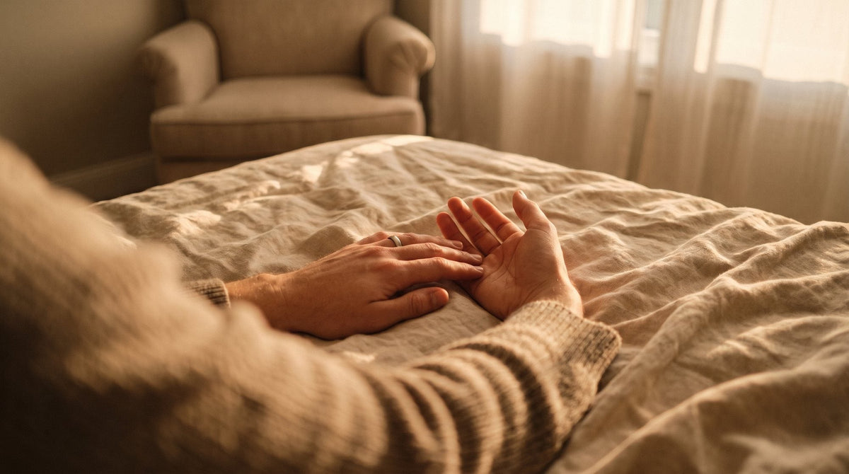Deux mains au repos sur un drap de lin couleur sable, dans une lumière dorée, devant un fauteuil vide tourné vers le lit
