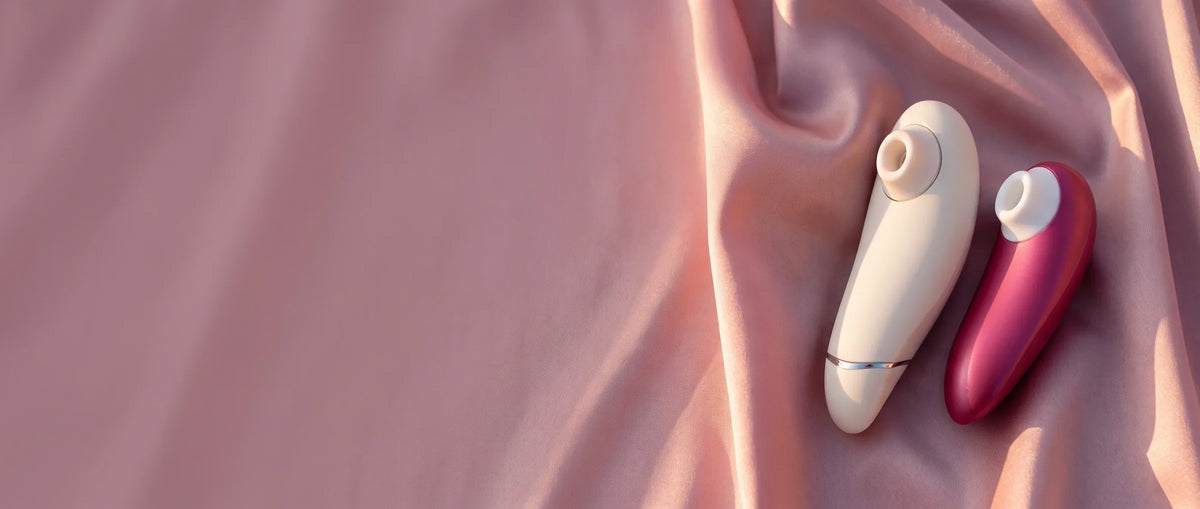 Deux stimulateurs Womanizer posés sur du satin blush, zone claire à gauche.