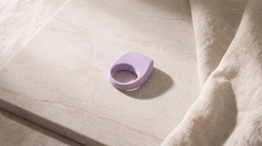 Anneau pénien vibrant LELO Tor 3 lilas posé sur une dalle de pierre claire veinée, à côté d’un pan de lin écru froissé