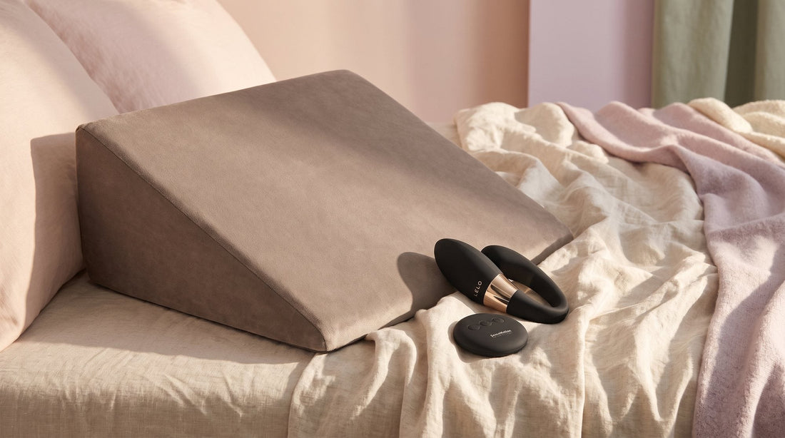 Coussin en coin à housse de velours taupe sur des draps de lin écru, à côté du LELO Tiani Duo noir et de sa télécommande