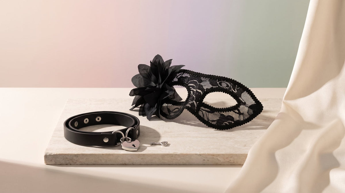 Masque vénitien en dentelle noire à fleur et collier noir cœur et clé posés sur une pierre claire, près d’un pan de satin crème