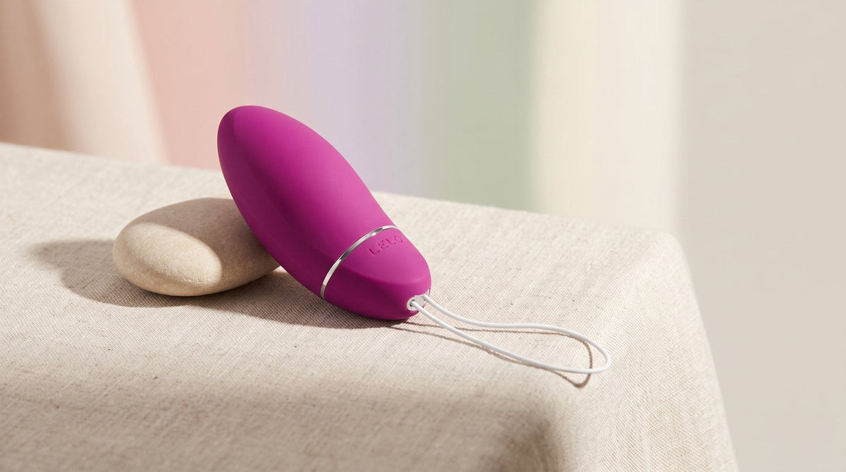Bola de Kegel LELO Luna Smart Bead sobre un lino natural de color crudo, junto a un canto rodado claro, con luz suave de mañana