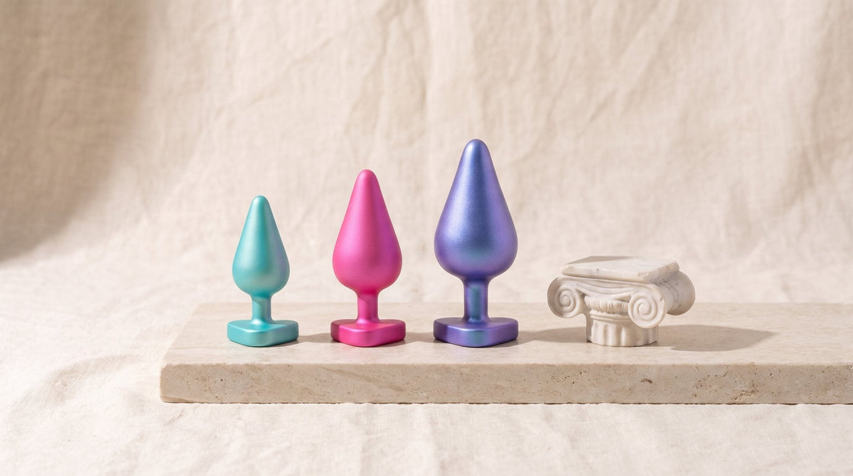 Tres plugs anales de silicona turquesa, rosa y azul violáceo, con base de corazón, junto a un capitel jónico de mármol