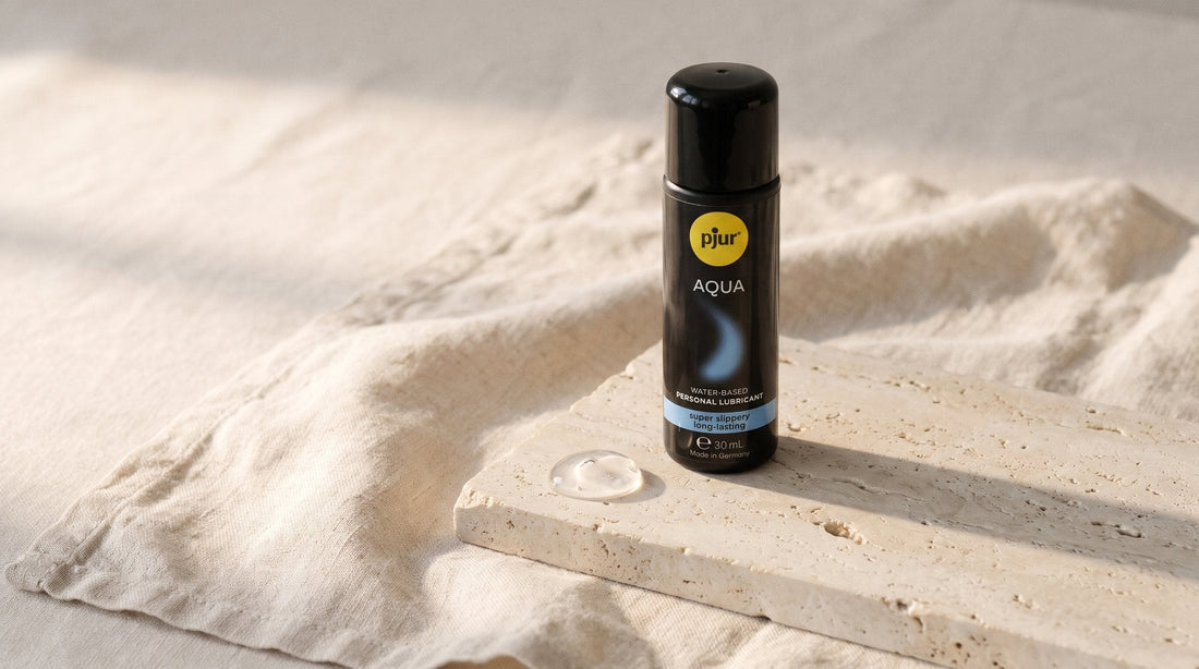 Frasco de lubricante pjur AQUA de pie sobre una losa de piedra clara y un lino crudo, junto a una gota de gel transparente