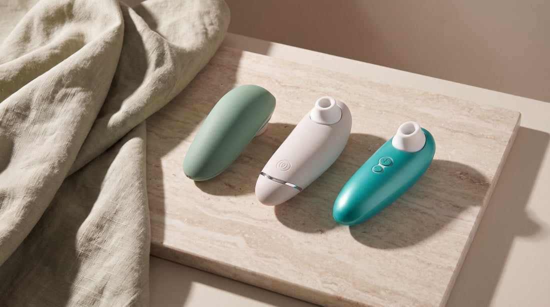 Trois stimulateurs Womanizer, Liberty 2 capot fermé, Premium 2 et Starlet 3, sur une dalle de pierre claire près d’un lin froissé
