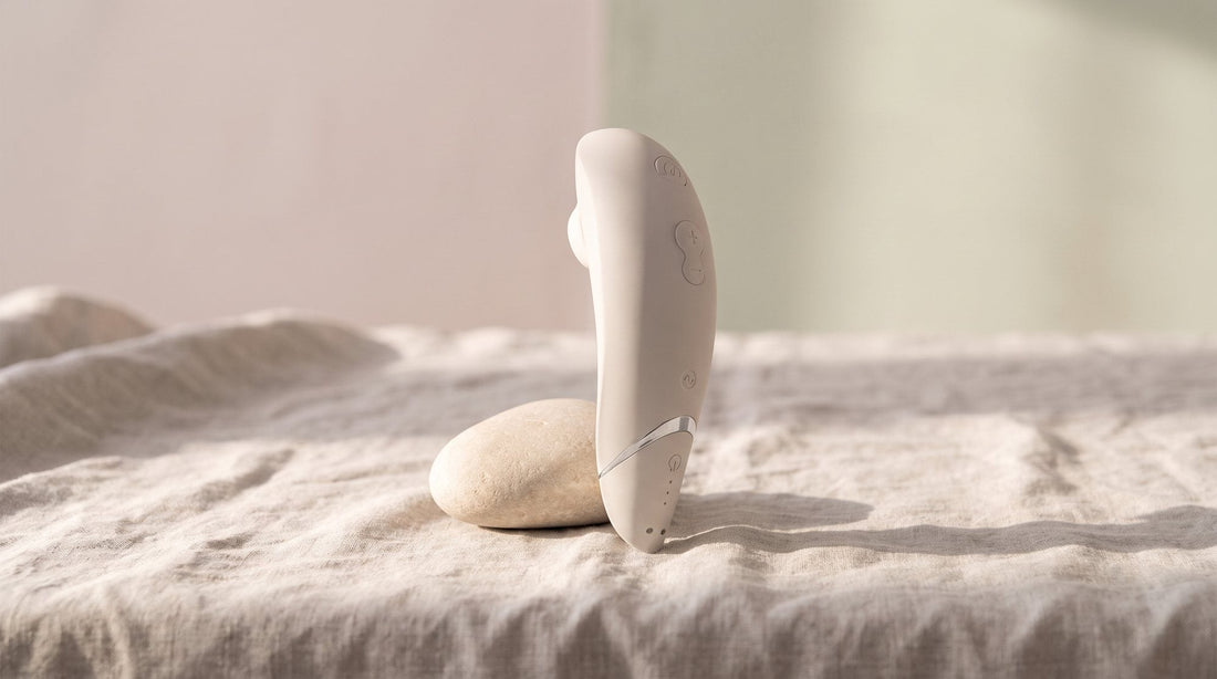 Stimulateur clitoridien Womanizer Premium 2 gris chaud posé debout sur une nappe de lin froissée, à côté d’un galet clair