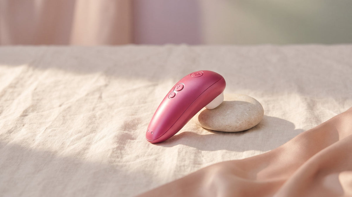 Womanizer Starlet 3 in Rosa auf zerknittertem Naturleinen, daneben ein heller Kieselstein und hautfarbener Satin im weichen Licht