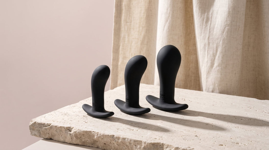 Drei Analplugs FUN FACTORY Bootie in S, M und L aus Silikon, aufgereiht auf hellem Kalkstein vor Leinenstoff im Seitenlicht