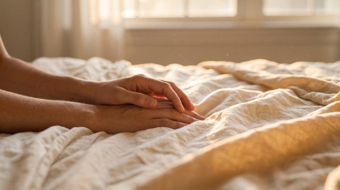 Deux mains de partenaires posées l’une sur l’autre sur des draps de lin froissés, dans une lumière dorée de fin de journée