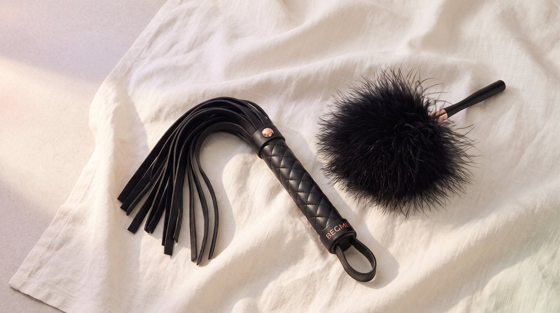 Schwarzer Flogger aus veganem Leder mit fächerartig ausgebreiteten Riemen neben einem schwarzen Federkitzler auf hellem Leinen