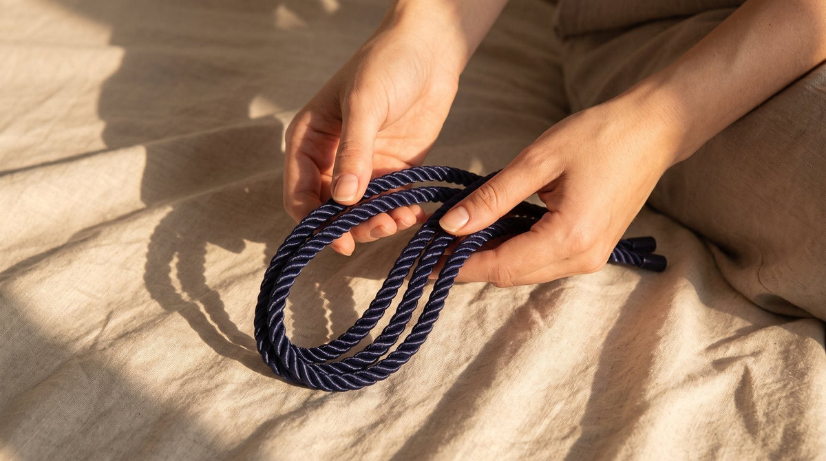 Zwei Hände legen ein blaues Nylonseil doppelt, über einem zerknitterten, ungebleichten Leinenlaken im warmen Licht des späten Nachmittags