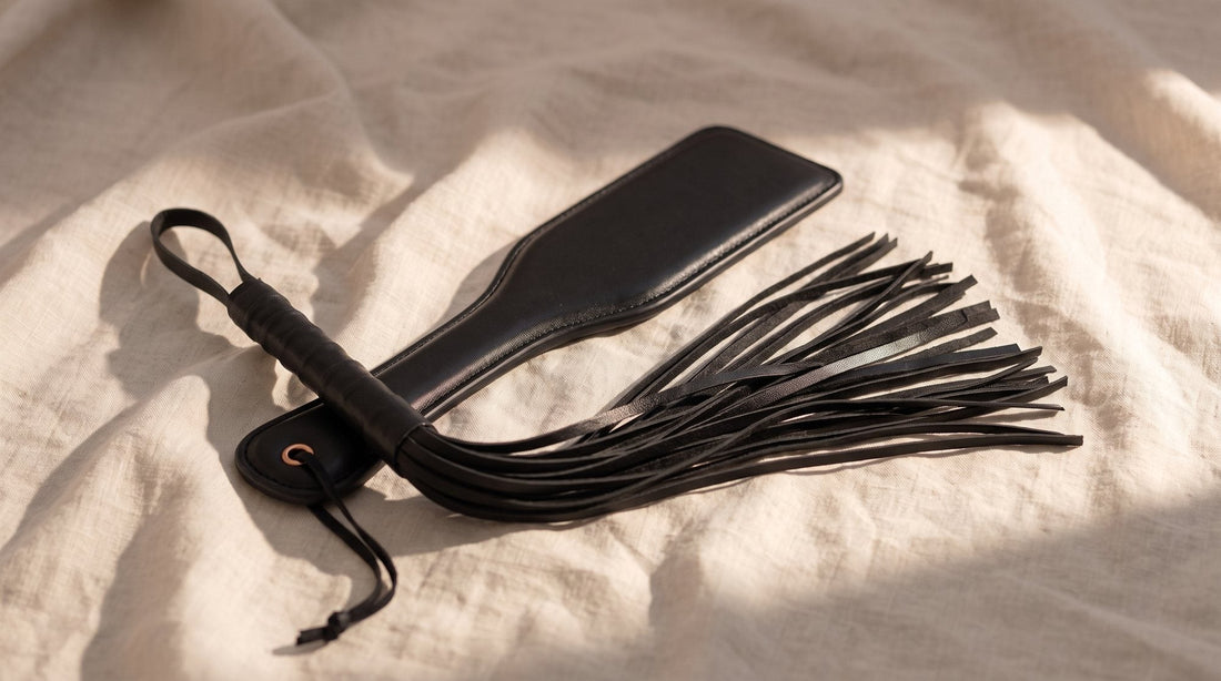 Schwarzes Paddle aus veganem Leder und schwarzer Flogger mit aufgefächerten Riemen auf zerknittertem, naturfarbenem Leinen