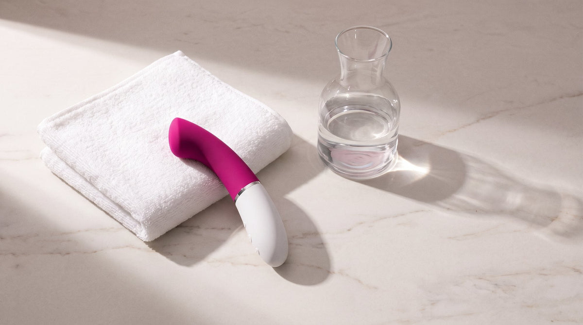 Vibrador LELO Gigi 3 rosa sobre una piedra clara veteada, junto a una toalla blanca doblada y una jarra de cristal con agua
