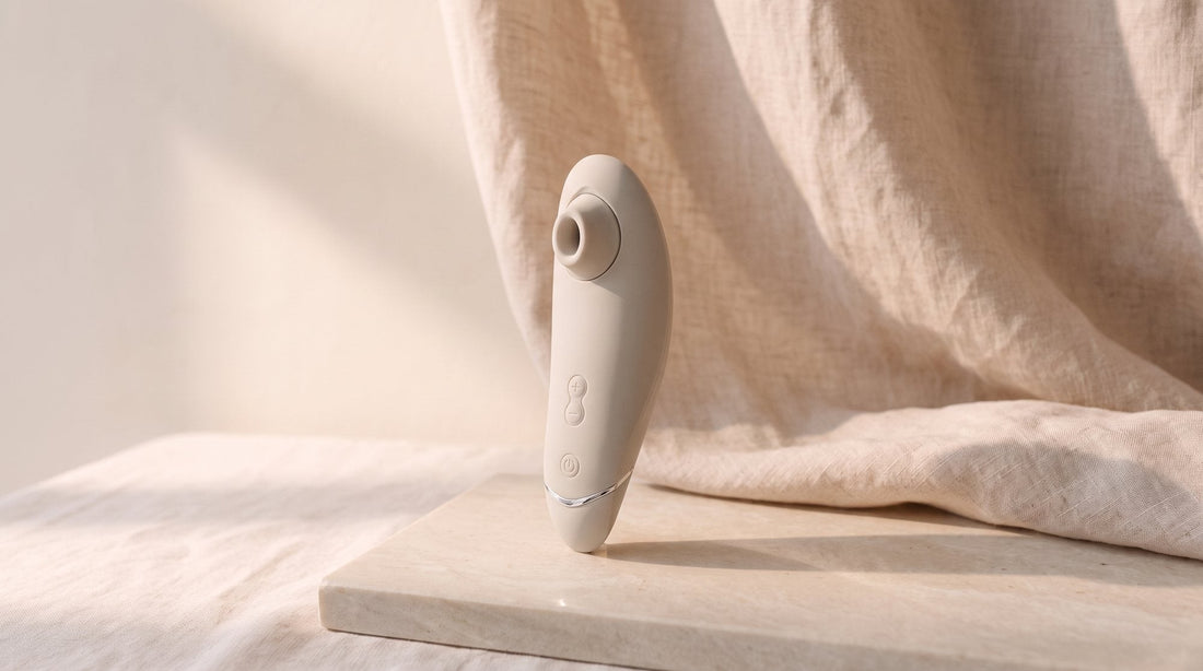 Druckwellenvibrator Womanizer Premium 2 in warmem Grau, aufrecht auf einer hellen Steinplatte vor zerknittertem Leinen, weiches Licht