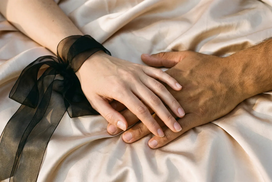 Une main de femme et une main d'homme posées l'une sur l'autre sur du satin champagne, ruban d'organza au poignet.