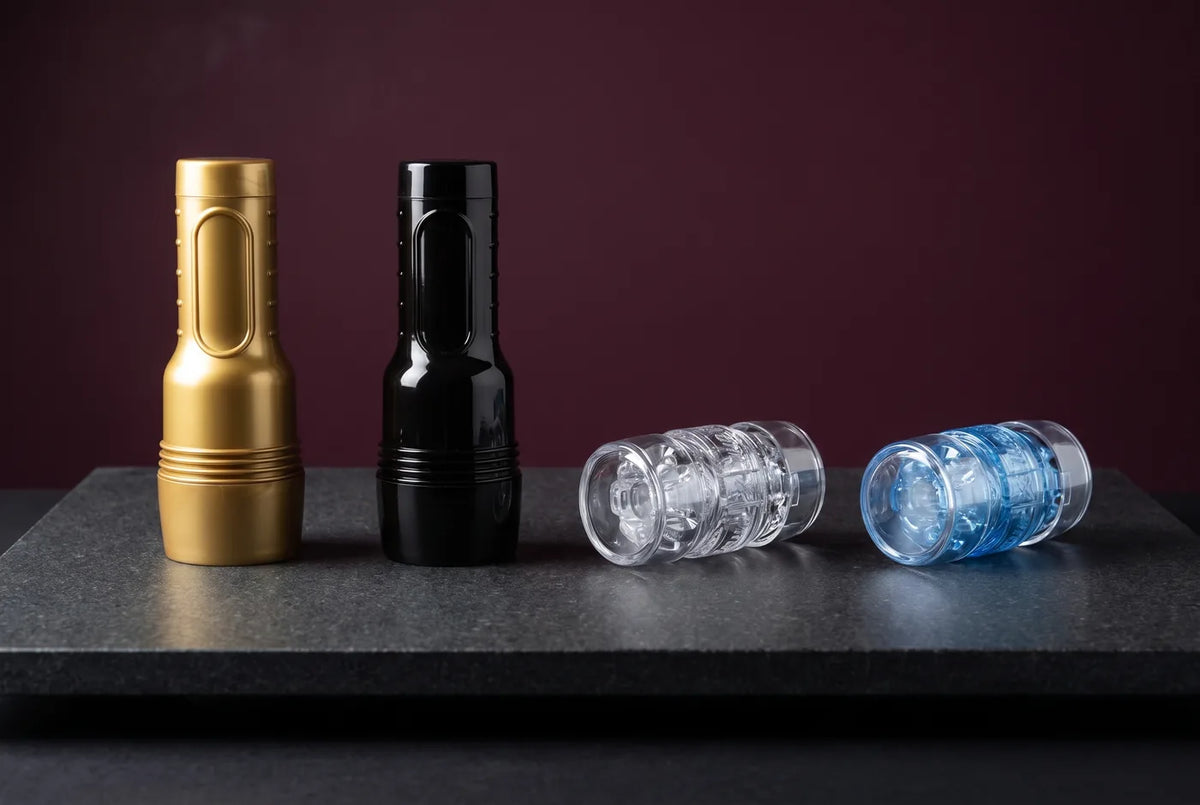 Vier Fleshlight-Masturbatoren mit aufgesetzten Kappen auf graphitgrauem Stein: GO Stamina Training Unit in Gold, GO Surge in Schwarz, Quickshot Vantage transparent und Quickshot Turbo in Blue Ice.