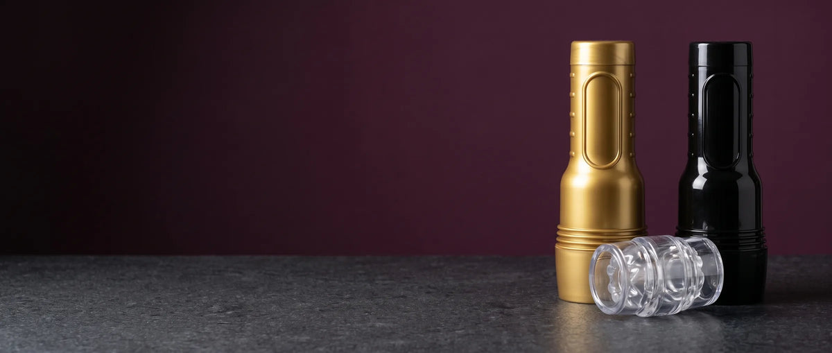Drei Fleshlight-Masturbatoren in geschlossenen Gehäusen – GO in Gold, GO in Schwarz und der transparente Quickshot Vantage – auf graphitgrauem Stein vor pflaumenfarbener Wand.