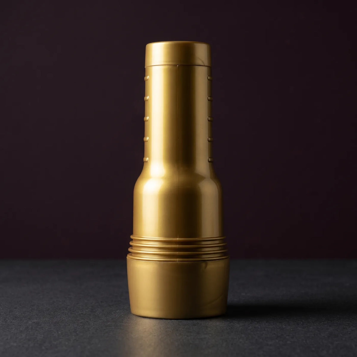 Fleshlight GO Stamina Training Unit Lady, kompakter Masturbator im goldenen Gehäuse, mit aufgesetzter Kappe stehend auf graphitgrauem Stein vor pflaumenfarbener Wand.