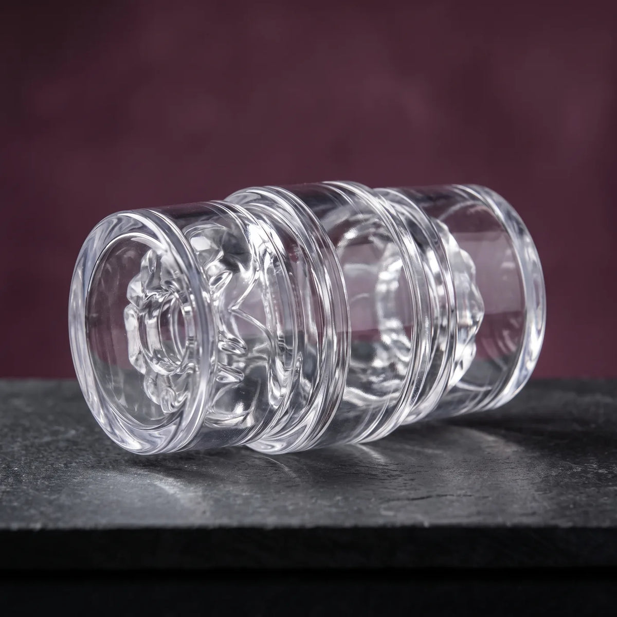 Fleshlight Quickshot Vantage, kompakter transparenter Masturbator mit beiden Kappen, auf graphitgrauem Stein vor pflaumenfarbener Wand.