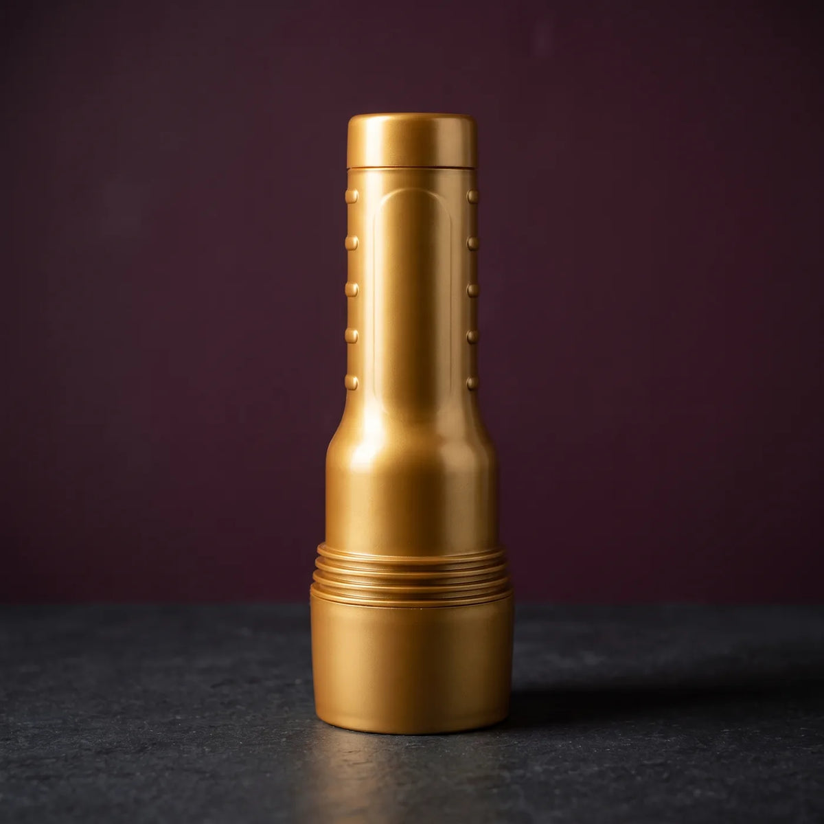 Fleshlight Stamina Training Unit, Masturbator im goldenen Gehäuse, mit aufgesetzter Kappe stehend auf graphitgrauem Stein vor pflaumenfarbener Wand.