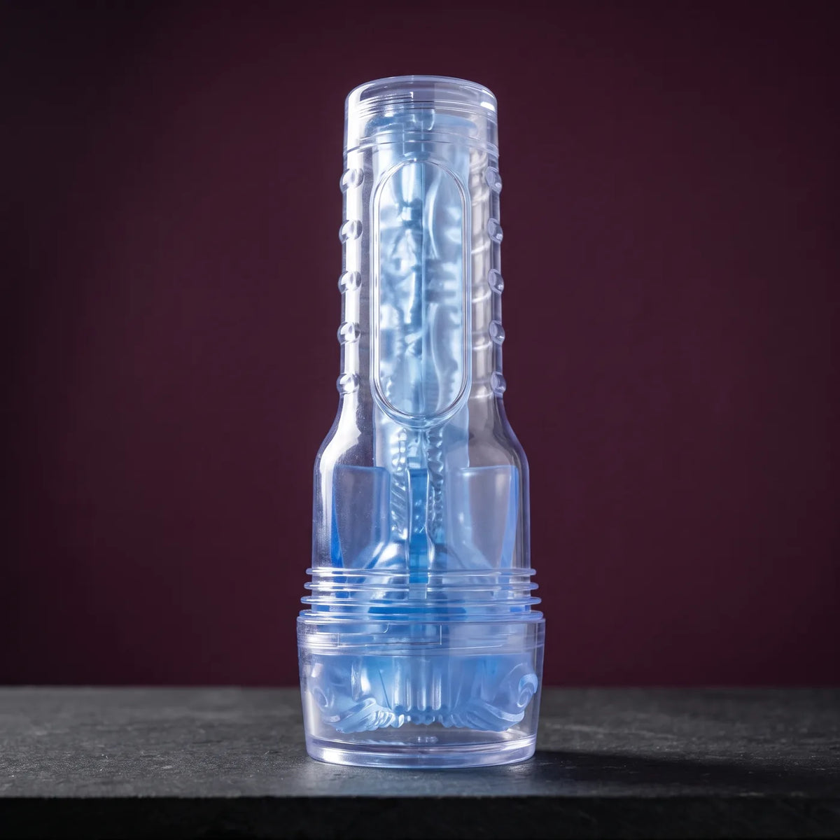 Fleshlight Turbo Thrust Blue Ice, Masturbator im transparent-blauen Gehäuse, mit aufgesetzter Kappe stehend auf graphitgrauem Stein vor pflaumenfarbener Wand.