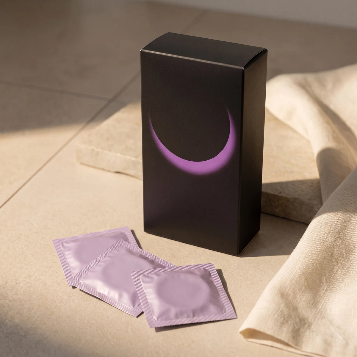 Schwarze Kondomschachtel mit violettem Bogen und einzelne unbedruckte Kondomverpackungen auf heller Steinplatte im Sonnenlicht.