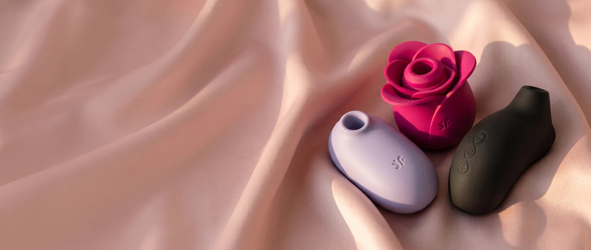 Tres succionadores de clítoris, Satisfyer Pro 2 Modern Blossom fucsia, Satisfyer Pro To Go 2 lila y LELO Sona negro, sobre satén rosa.
