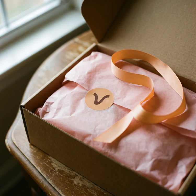 Papier de soie blush, ruban abricot et pastille ronde marquée d'un V.