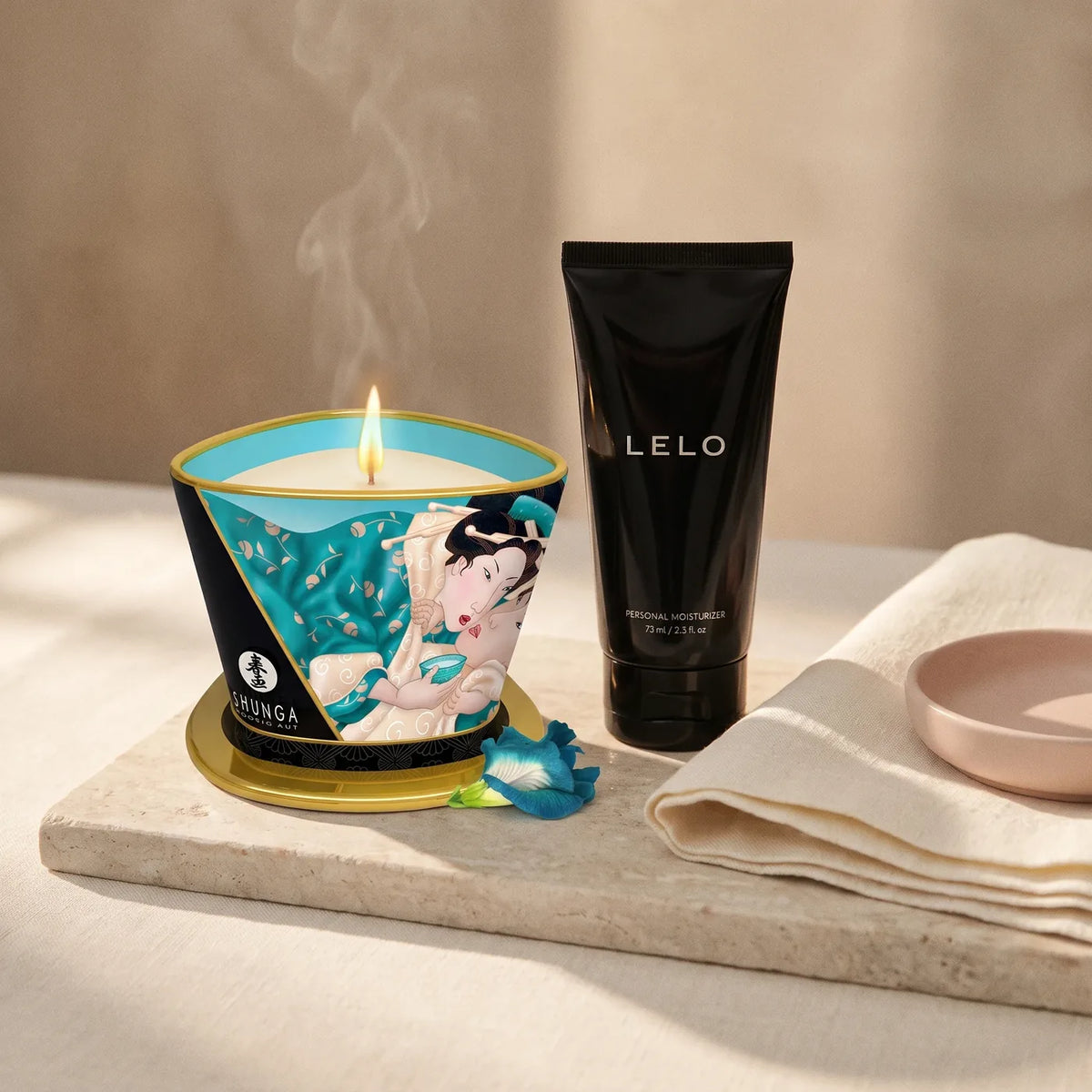 Bougie de massage allumée et flacon de gel sur pierre claire et lin ivoire.