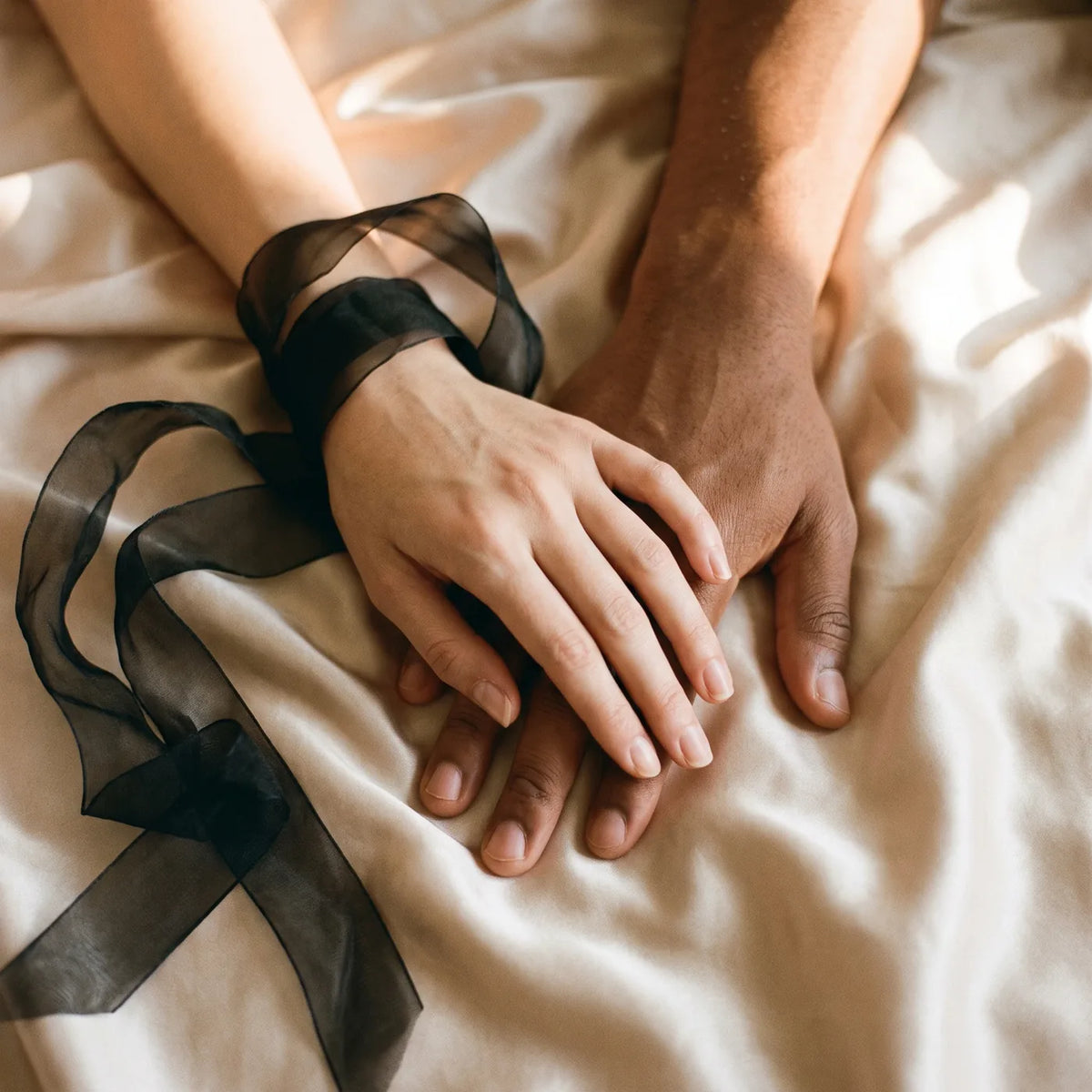 Une main de femme et une main d'homme posées l'une sur l'autre sur du satin champagne, ruban d'organza au poignet.