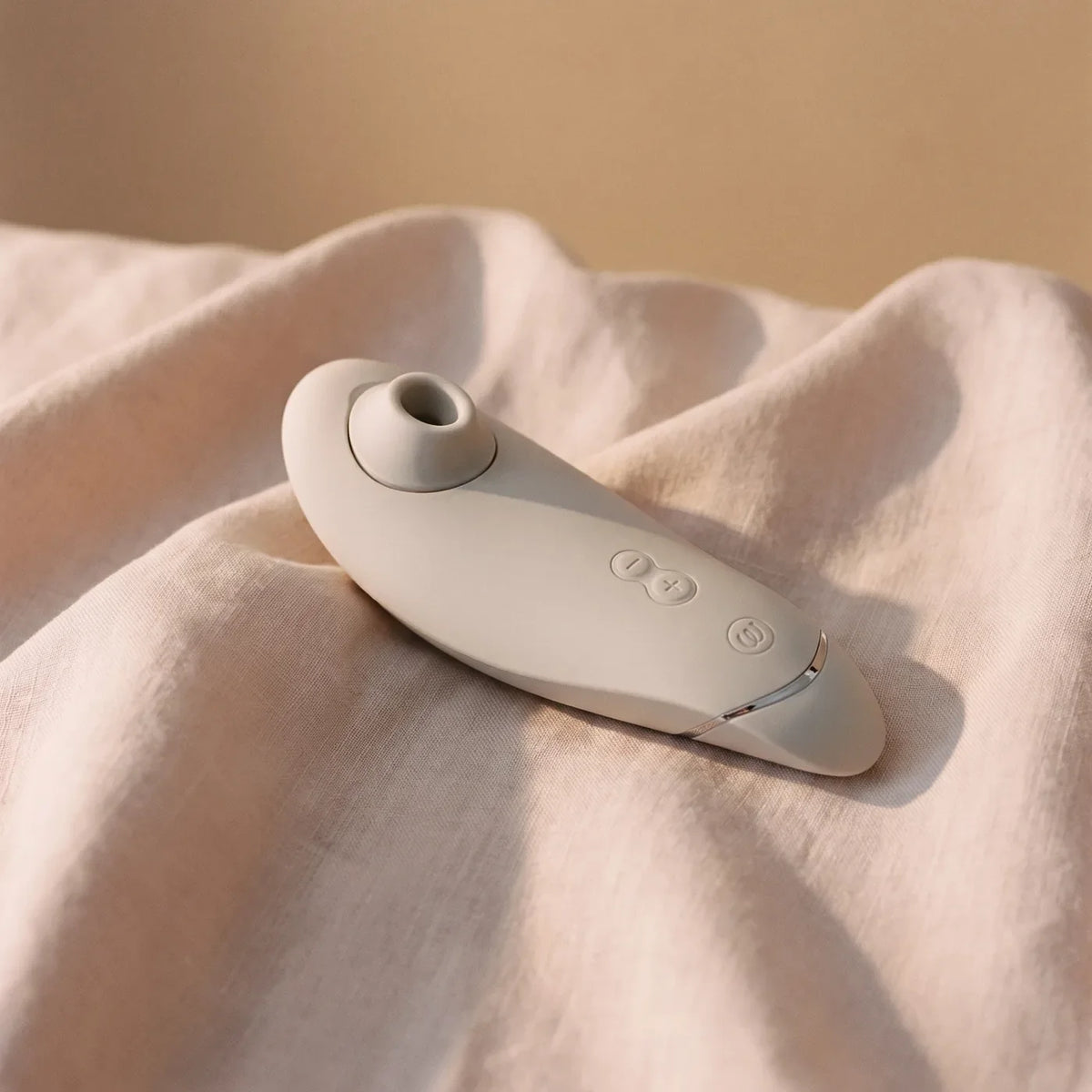 Womanizer Premium 2 gris chaud posé sur du lin ivoire.