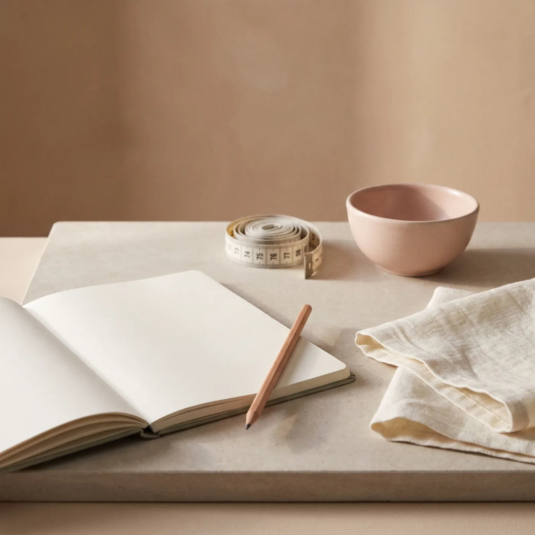 Carnet ouvert, mètre de couturière et serviette de lin pliée sur une tablette de pierre claire.
