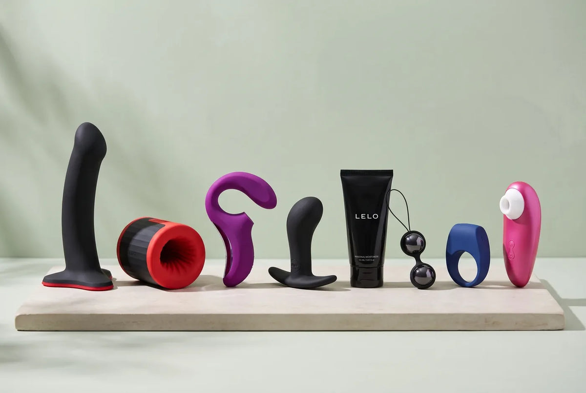 Huit objets de la sélection Voluptuo alignés sur une tablette de pierre claire : silicone souple, stimulateur à air, masseur, anneau, boules, flacon de soin.
